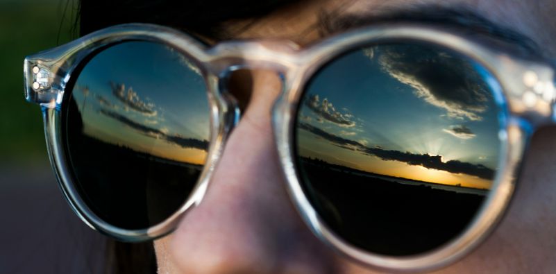 A close-up of a man wearing sunglasses, showing a reflection of the sun in the lenses