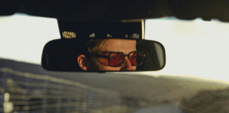 A man wearing sunglasses reflected in a car’s rear-view mirror.