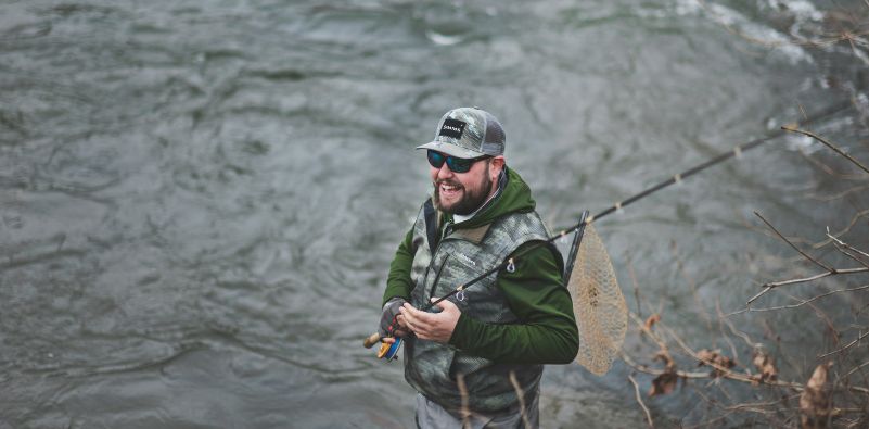A man in the middle of a river fishing, while wearing sunglasses and a cap